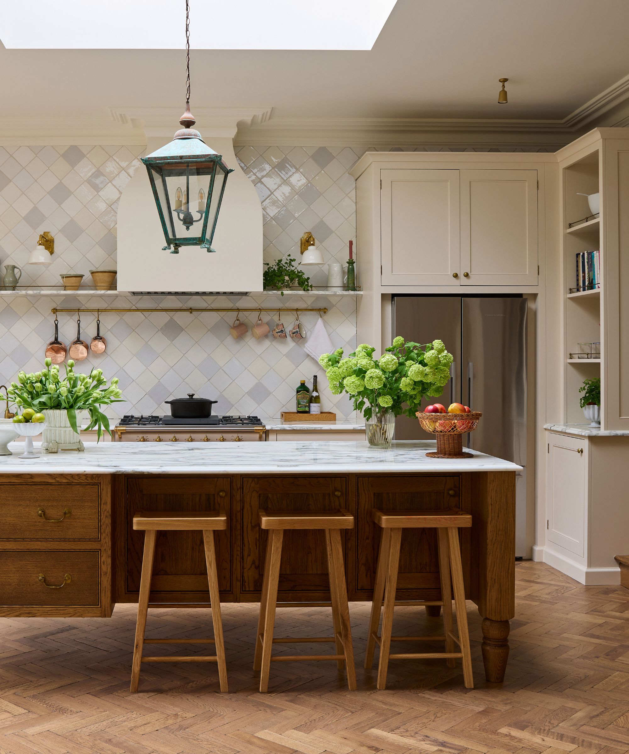 A classic English-style kitchen featuring a large oak island with a marble countertop, three wooden stools, and a distinctive verdigris lantern hanging above. The background shows cream cabinetry and a diamond-patterned tiled backsplash