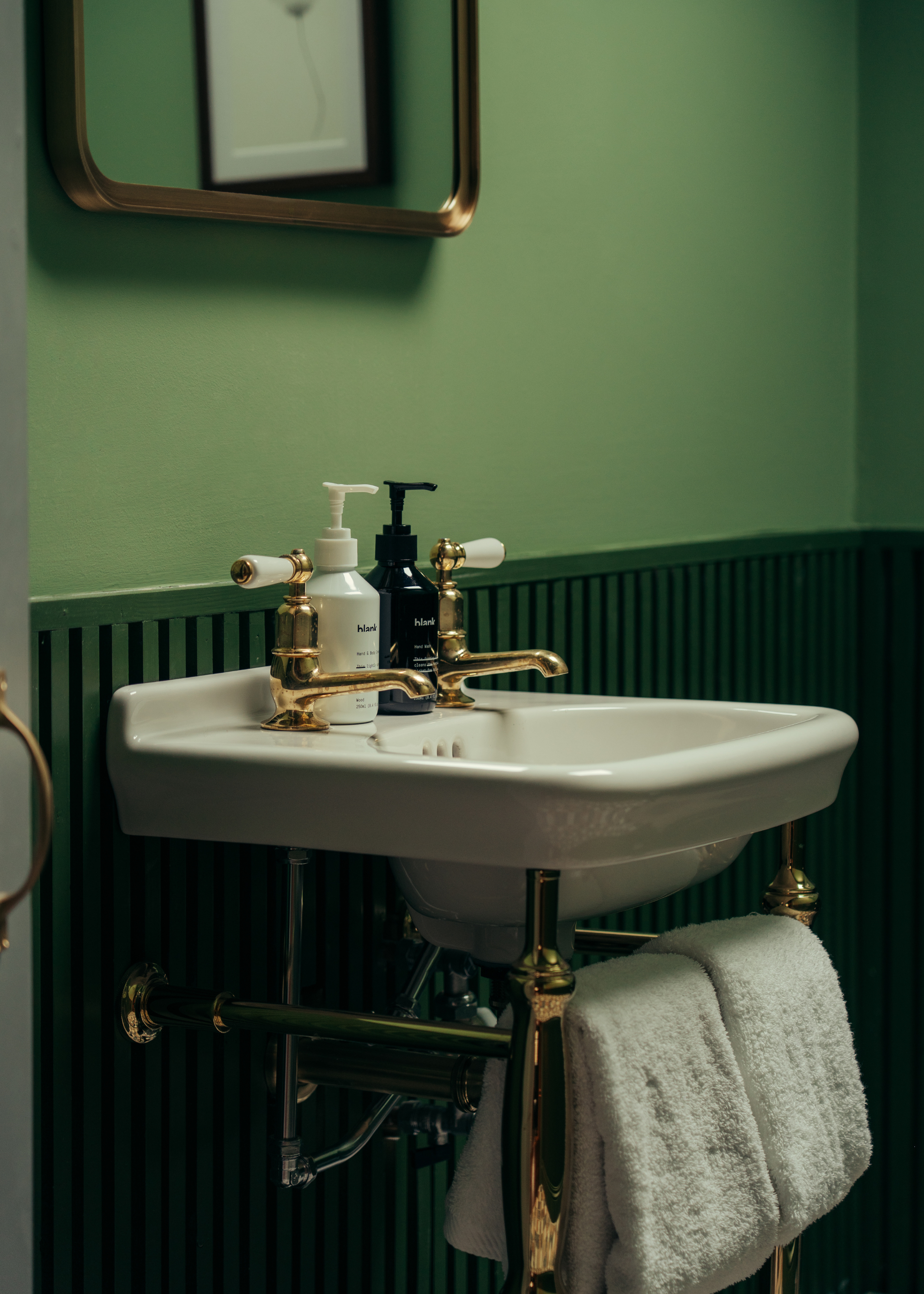 A green bathroom with a mounted mirror, a white sink, gold hardware, bath towels, and bottles of hand soap and moisturizer