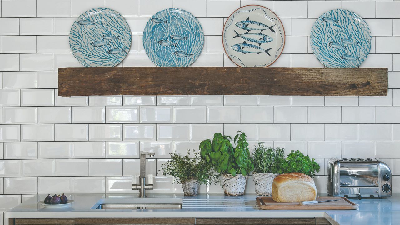 A white-tiled kitchen with potted herbs on the worktop and fish-motif plates displayed on a rustic shelf