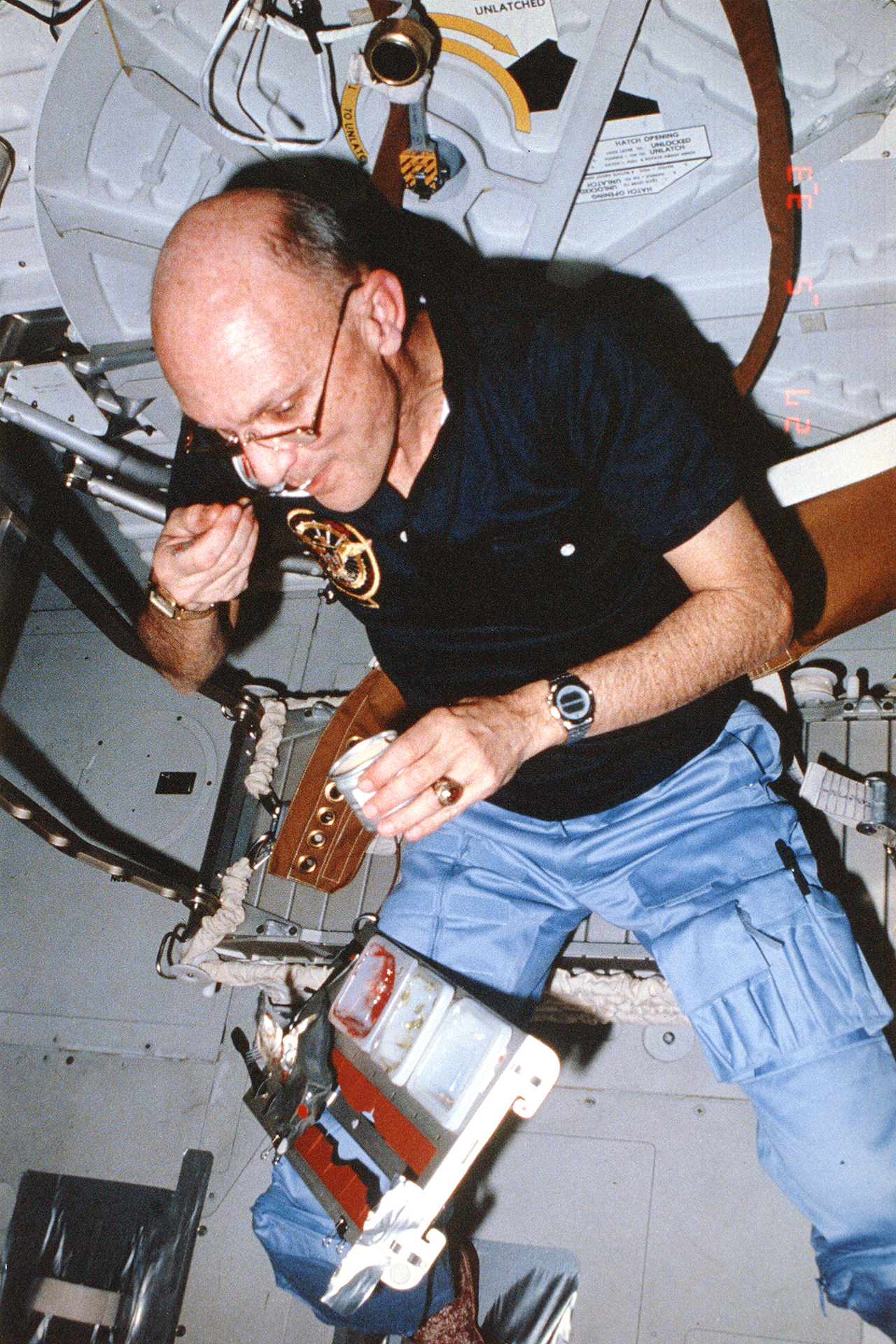 a man in a blue shirt eats while floating inside a white-walled spacecraft.