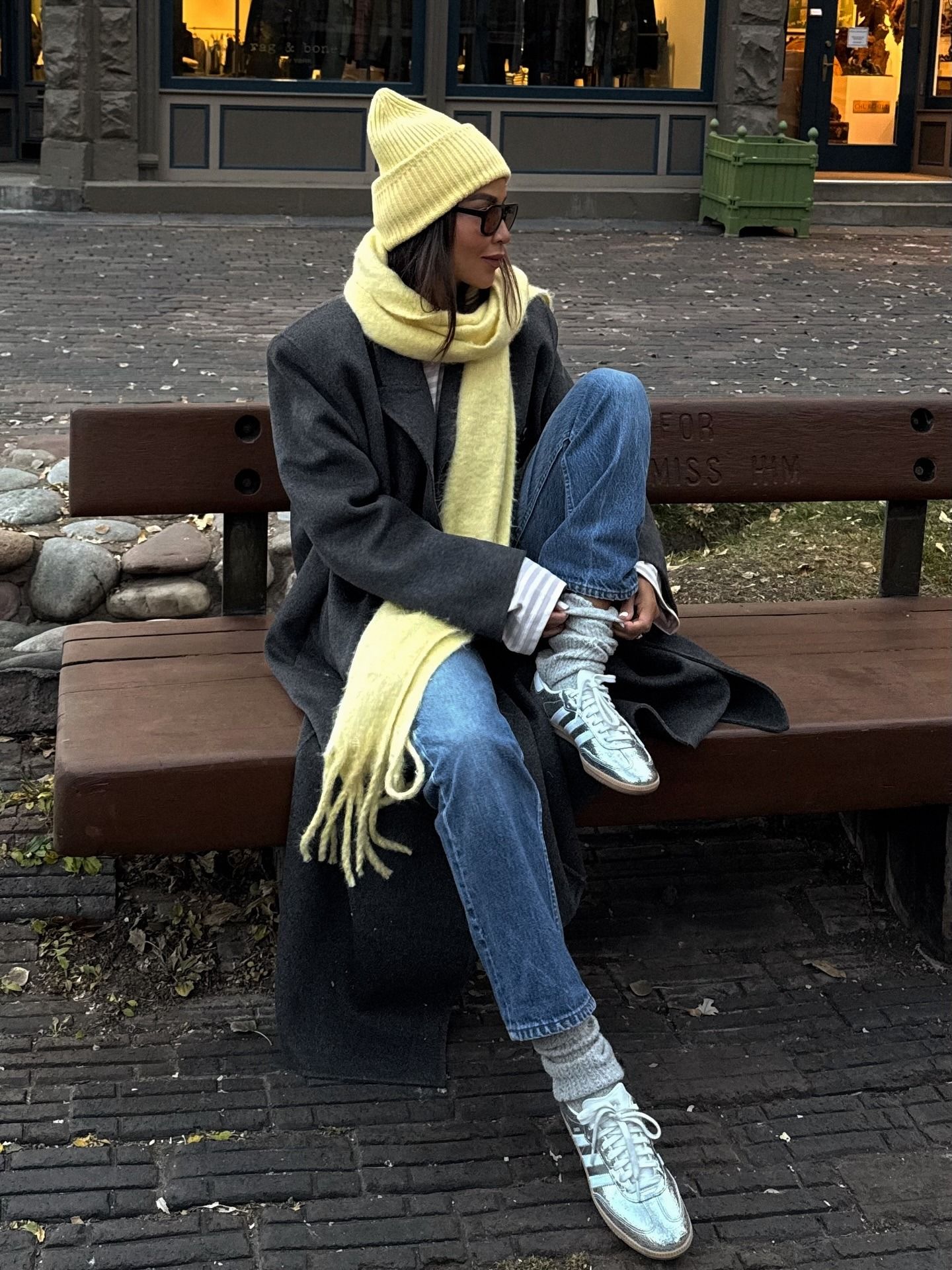 Fashion person Julie Sariñana @sincerelyjules wears a leather trainer outfit. She wears a metallic trainer, jeans long coat and butter yellow hat and scarf.