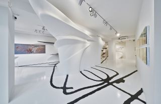 Galerie Gmurzynska Gallery. The room is all white, with black lines on the floor. At the center, is an asymmetrical white structure, that's permanent in the gallery. There is an abstract painting to the left.