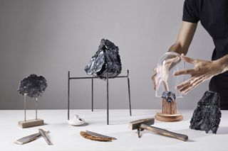 An image showcasing multiple artistic items placed on a white table. Black sculptural stone like objects, wooden straight pin hammer. A man in black short sleeve tshirt holder a glass dome over a black sculptured stone placed on a wooden base