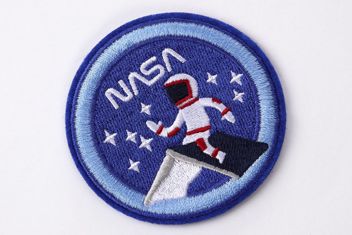 Embroidered patch from the NASA-inspired Limited Edition Notebooks from Moleskine. It's a round dark blue patch with a light blue border. The word "NASA" is at the top in white. A person in a white astronaut suit with red trim is sitting on the roof of a house. They are surrounded by white stars.