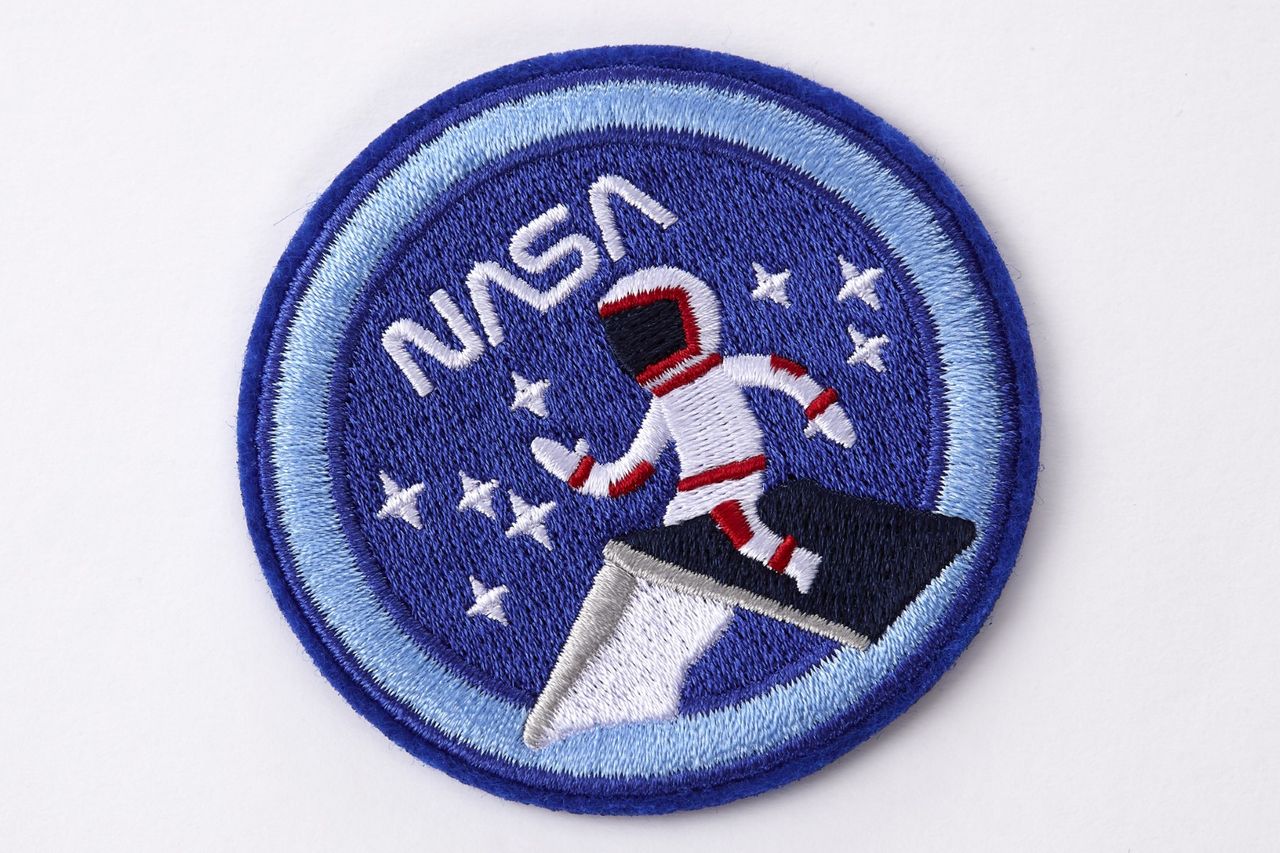 Embroidered patch from the NASA-inspired Limited Edition Notebooks from Moleskine. It's a round dark blue patch with a light blue border. The word "NASA" is at the top in white. A person in a white astronaut suit with red trim is sitting on the roof of a house. They are surrounded by white stars.