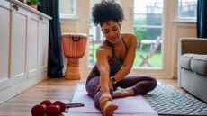 A woman sits on an exercise mat at home, stretching one leg in front with the other leg crossed in front of her. Behind her, we see a drum and some dumbbells, along with large glass doors leading to a grassy backyard.