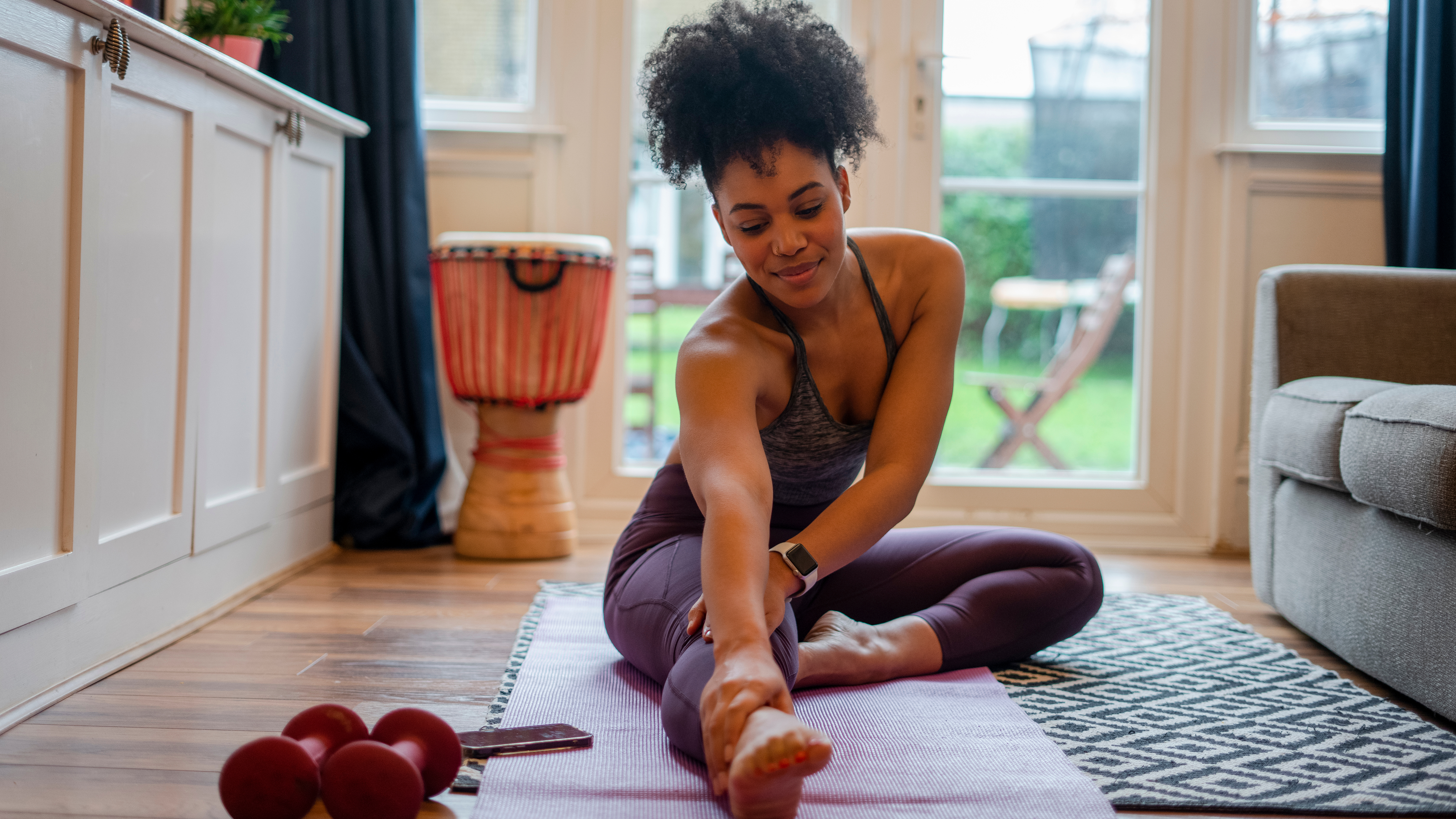 A woman sits on an exercise mat at home, stretching one leg in front with the other leg crossed in front of her. Behind her, we see a drum and some dumbbells, along with large glass doors leading to a grassy backyard.