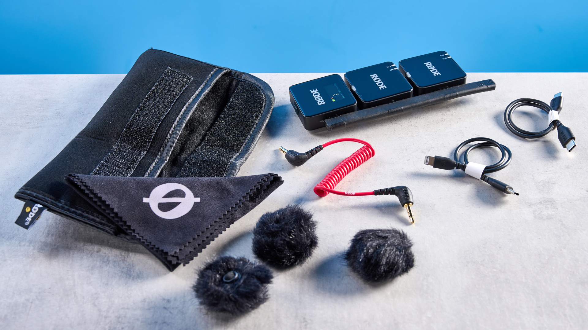 the rode wireless go gen 3 microphone with one receiver, two transmitters, shown against a blue background and with accessories like dead cat, charging ports, and cables