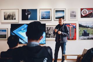 Man talking to an audience beside flat screen television with photos in the background