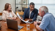 Advisor Explaining The Details Of The Contract He Is Holding In His Hand To His Mature Adult Clients Around Their Kitchen Table