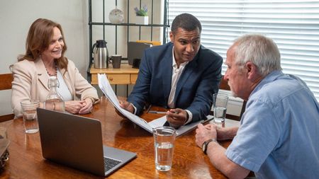 Advisor Explaining The Details Of The Contract He Is Holding In His Hand To His Mature Adult Clients Around Their Kitchen Table