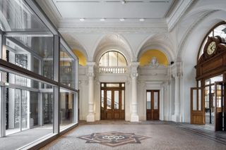 The entry to The Bourse de Commerce in Paris. To the left are large glass doors. Marble floors, white walls with ornaments above the doors, and heavy wooden doors decorate this spacious entry hall.