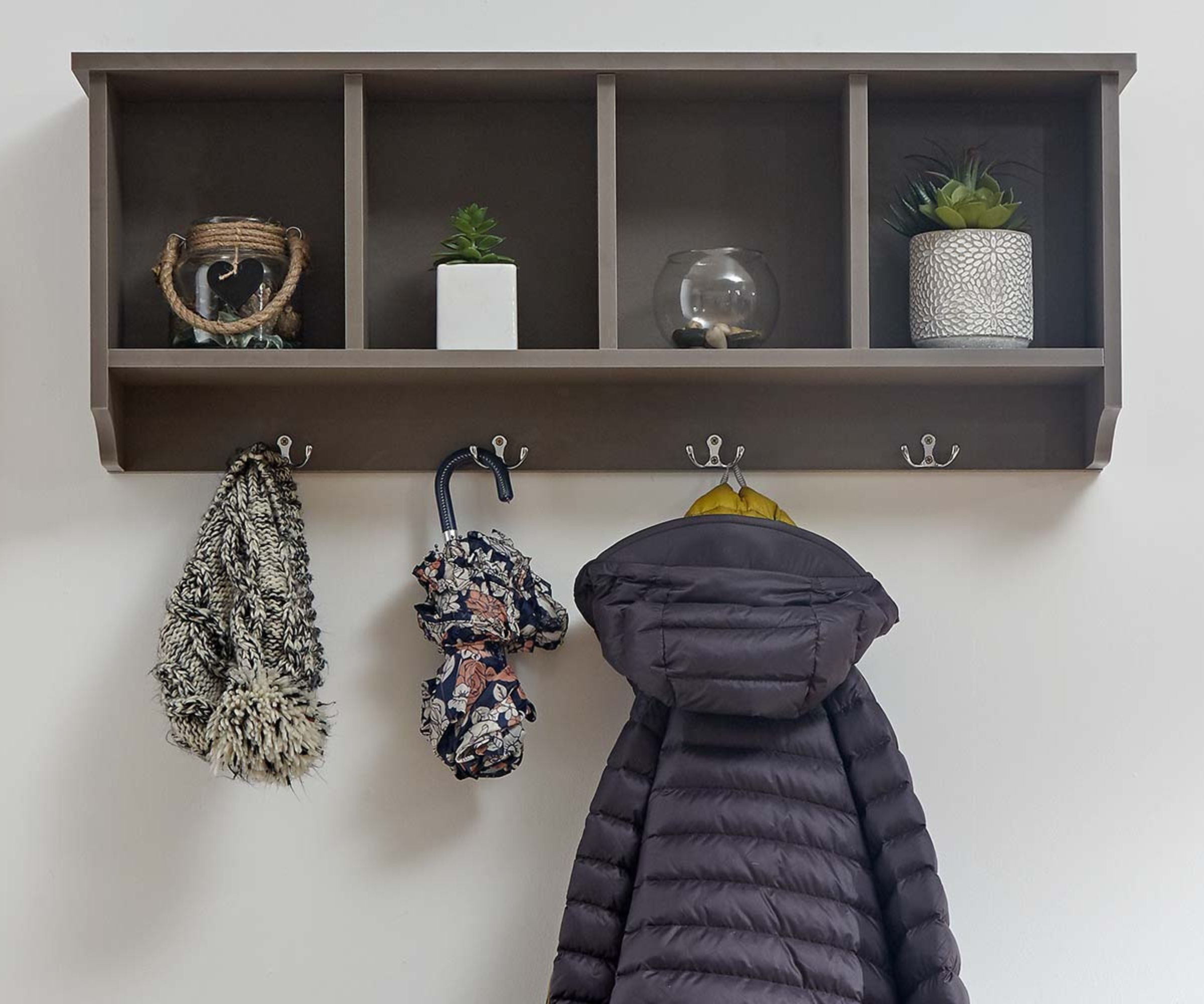 A dark grey wall-mounted hallway shelf unit with four cubby holes and four chrome double hooks, holding a puffer jacket, scarf and umbrella.
