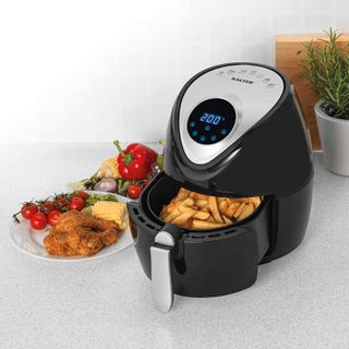 A black Salter branded air fryer containing fries, with plate of fried chicken, corn on the cob and vine tomato garnish