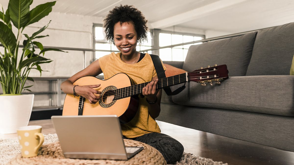 Woman plays acoustic guitar sat on a sofa in front of a laptop