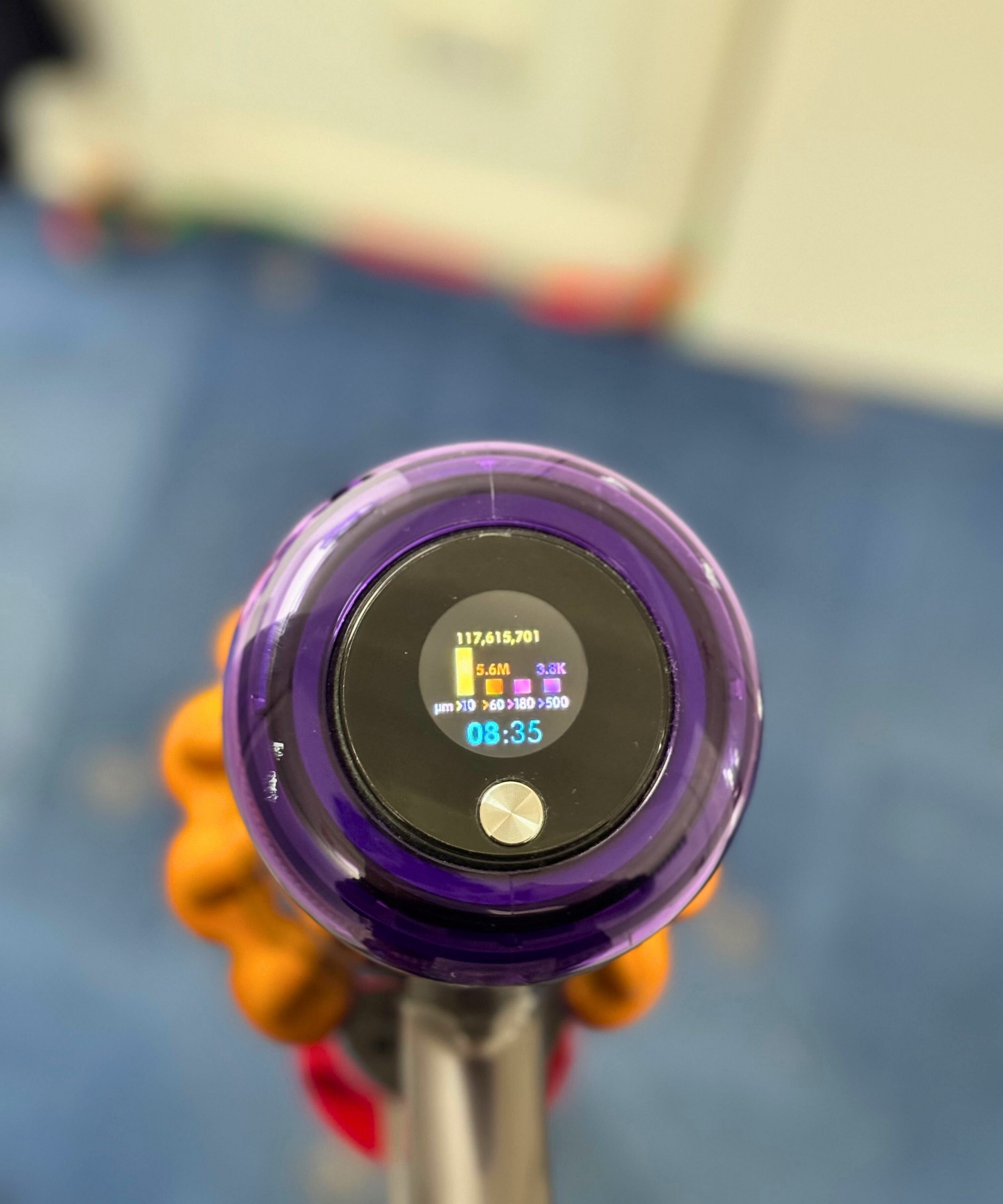 Closeup of the Dyson V15 Detect cordless vacuum's LED screen showing millions of particles picked up, above blurred-out blue carpet and white walls.