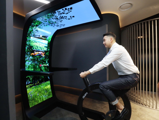 Man on fitness bike looking at LG Display Virtual ride, an emcompassing screen structure featuring flexible LG OLED panels.