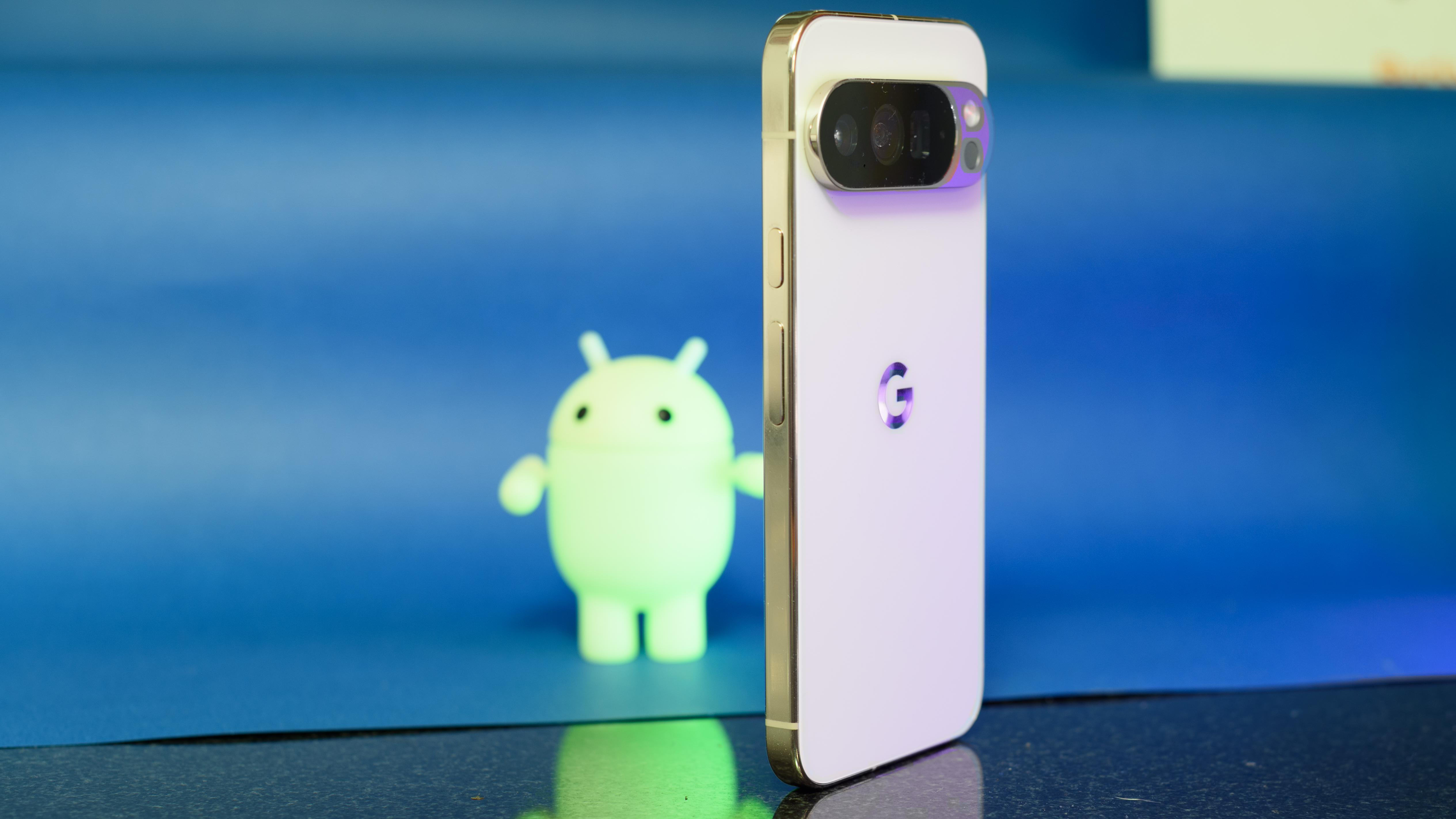 Google Pixel 10 Pro XL in Porcelain against a blue background with a green neon Android figurine in the background