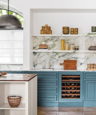 blue and white kitchen with marble floating shelves, kitchen island, stone flooring, wine cooler, chrome pendant light, baskets and vases on shelving,