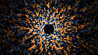 A black dot in the middle of a circle of blue, orange, and white smaller dots all on a black background