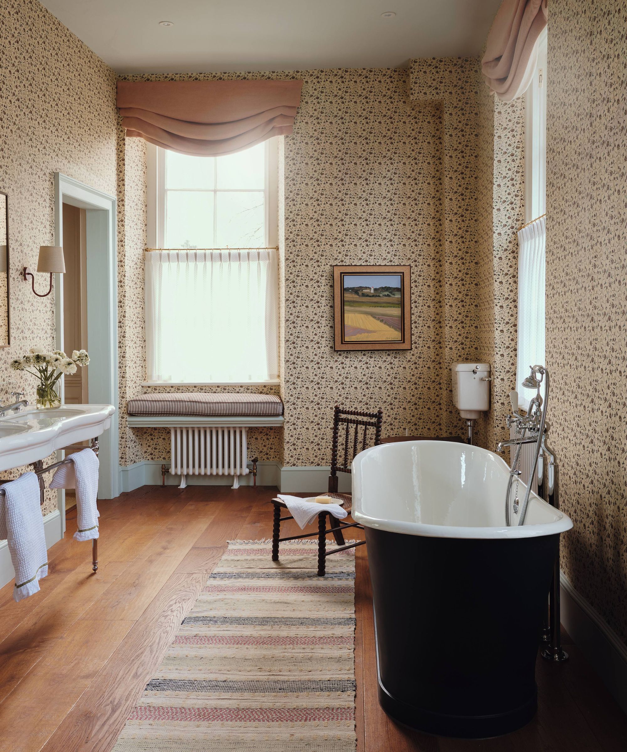 a ditsy print wallpapered bathroom with pink window blinds, cafe curtains, a large black freestanding tub, antique washstand and vintage chair and rug