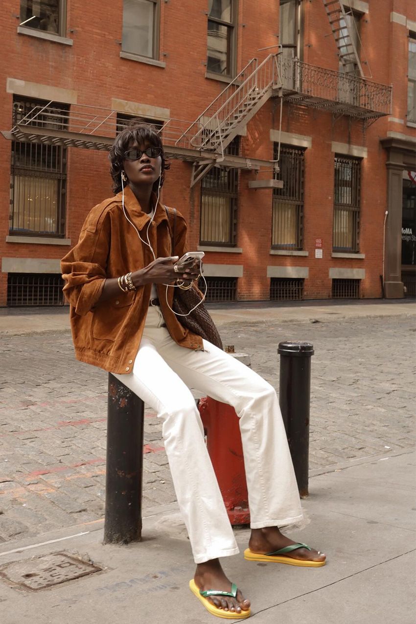 A woman sitting outside wearing a suede bomber jacket with a white shirt, white jeans, two-tone rubber flip-flops, gold jewelry, and black sunglasses.