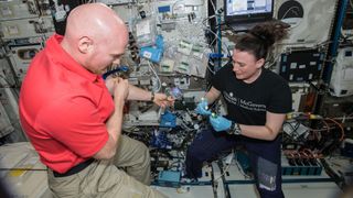 an astronaut floating in a cramped space station has his blood drawn by another astronaut