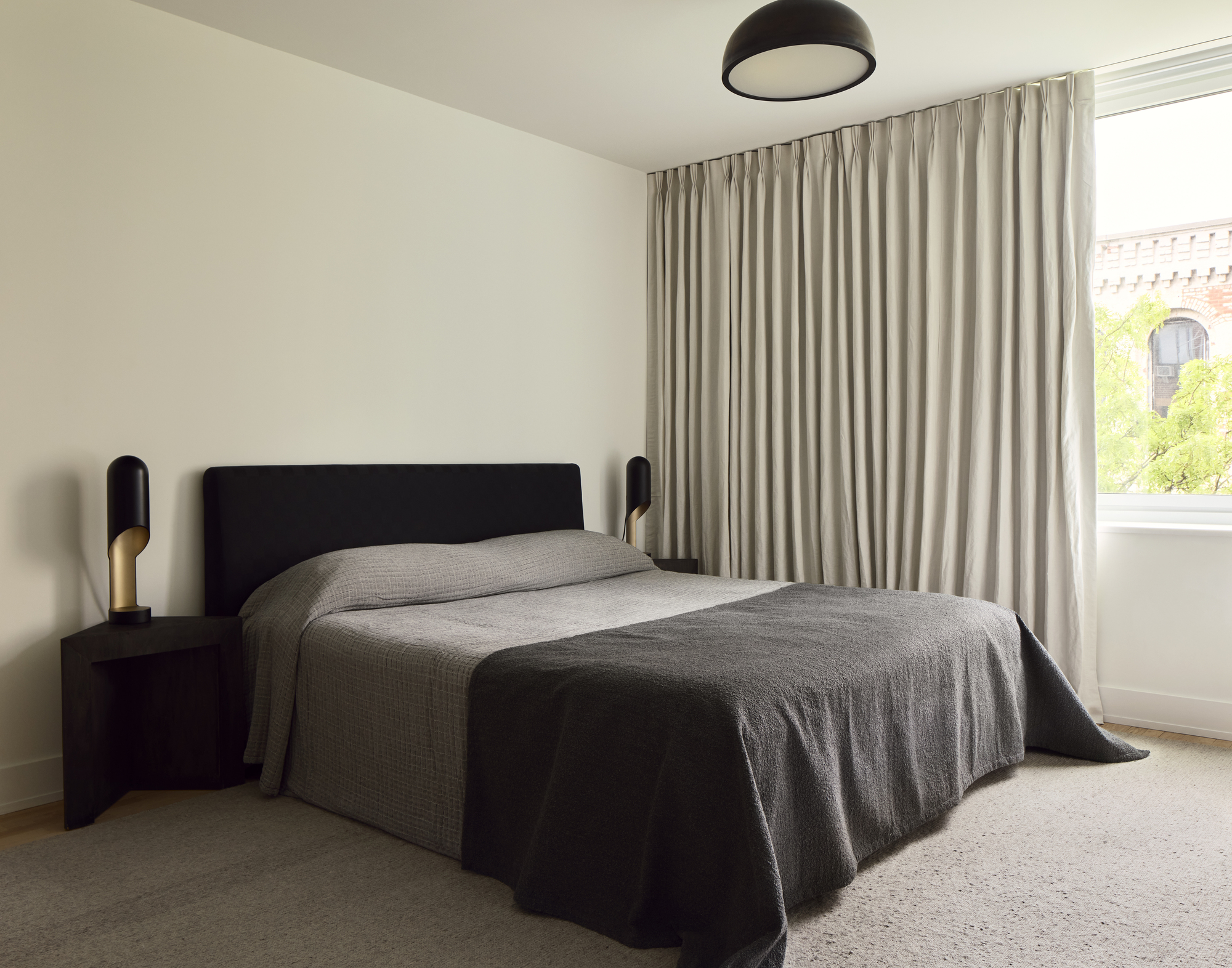 pared back bedroom with black headboard and throw, gray sheets, carpets, floor-to-ceiling drapes, and black wood side tables