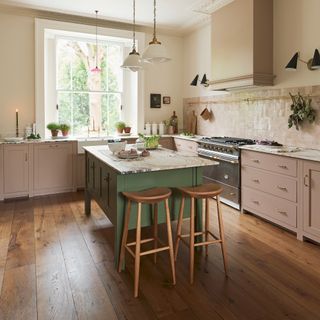 small u-shaped kitchen with cream walls and warm wood floor pink cabinetry and green island all with marble worktops with cat on the wooden stool