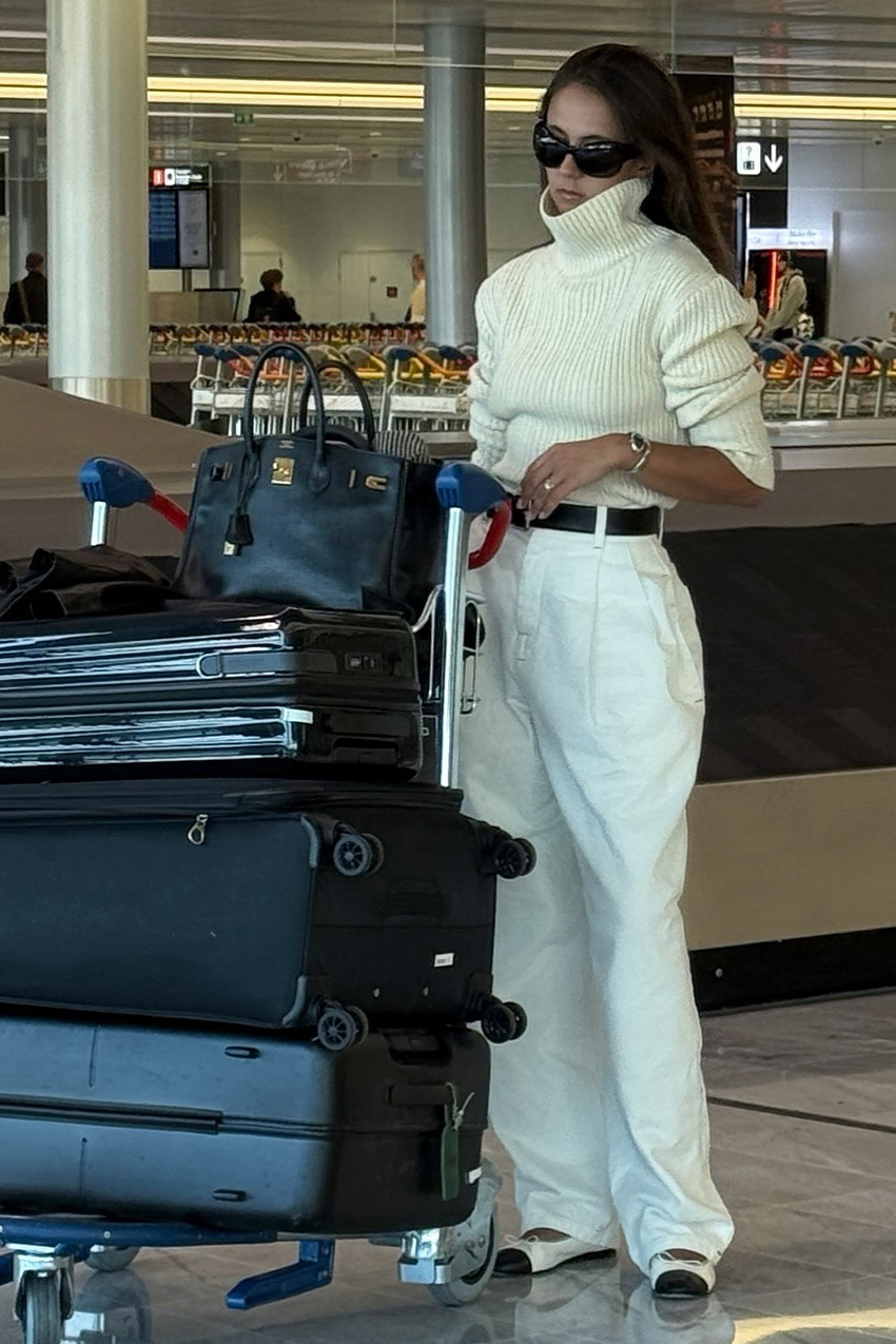 Shoe trends to wear with leggings, jeans, and skirts are shown in a street style photo of a woman standing in an airport terminal next to luggage, wearing black sunglasses, a white turtleneck sweater, a black belt, white baggy trousers, and black and white two-tone ballet flats.