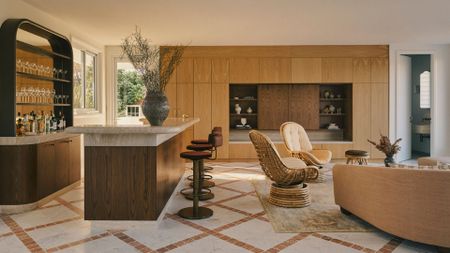 An open bar and living room with a mirrored bar, a stone and wood island, a set of swivel stools, a pair of woven chairs, and a TV console