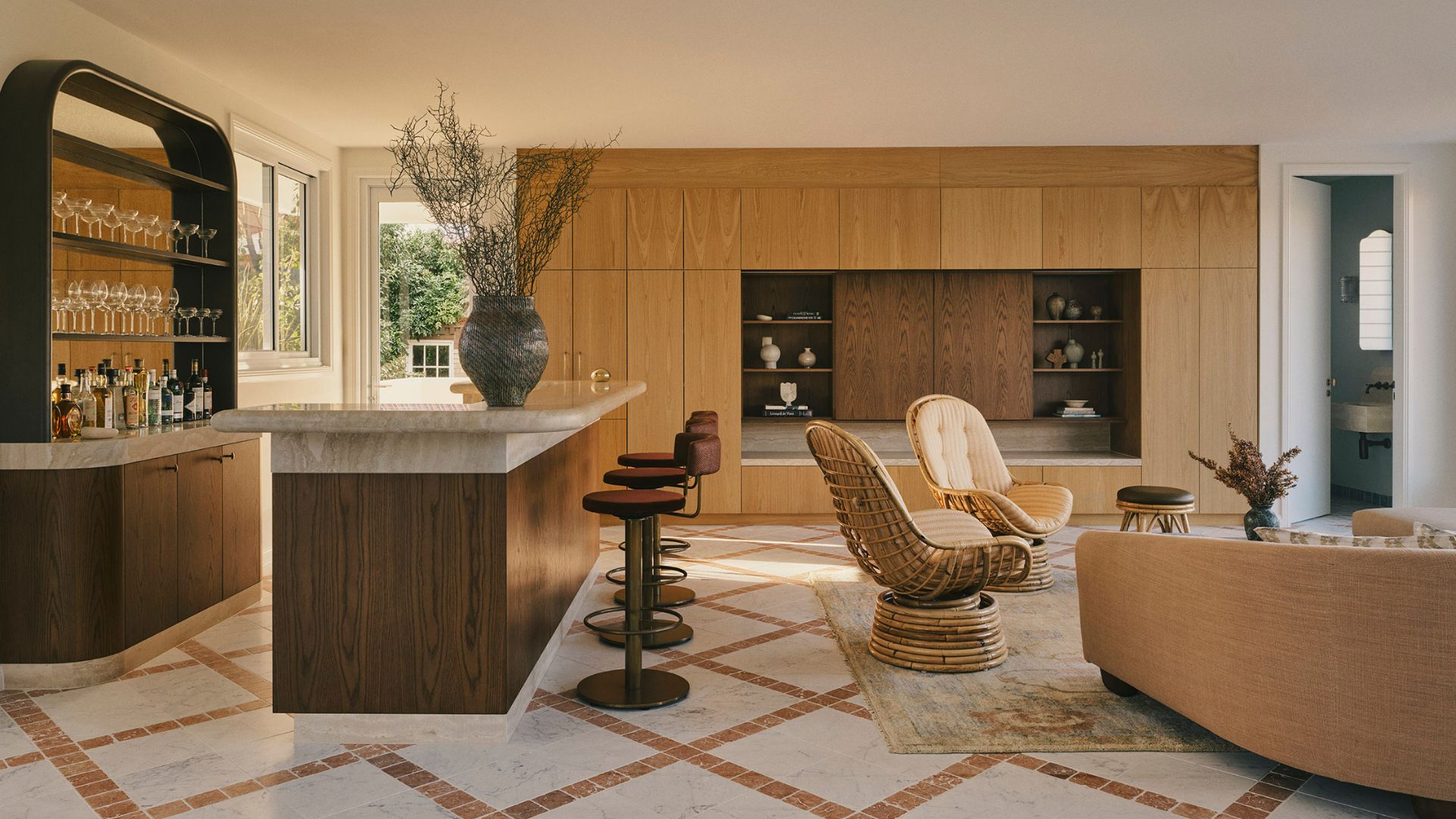 An open bar and living room with a mirrored bar, a stone and wood island, a set of swivel stools, a pair of woven chairs, and a TV console