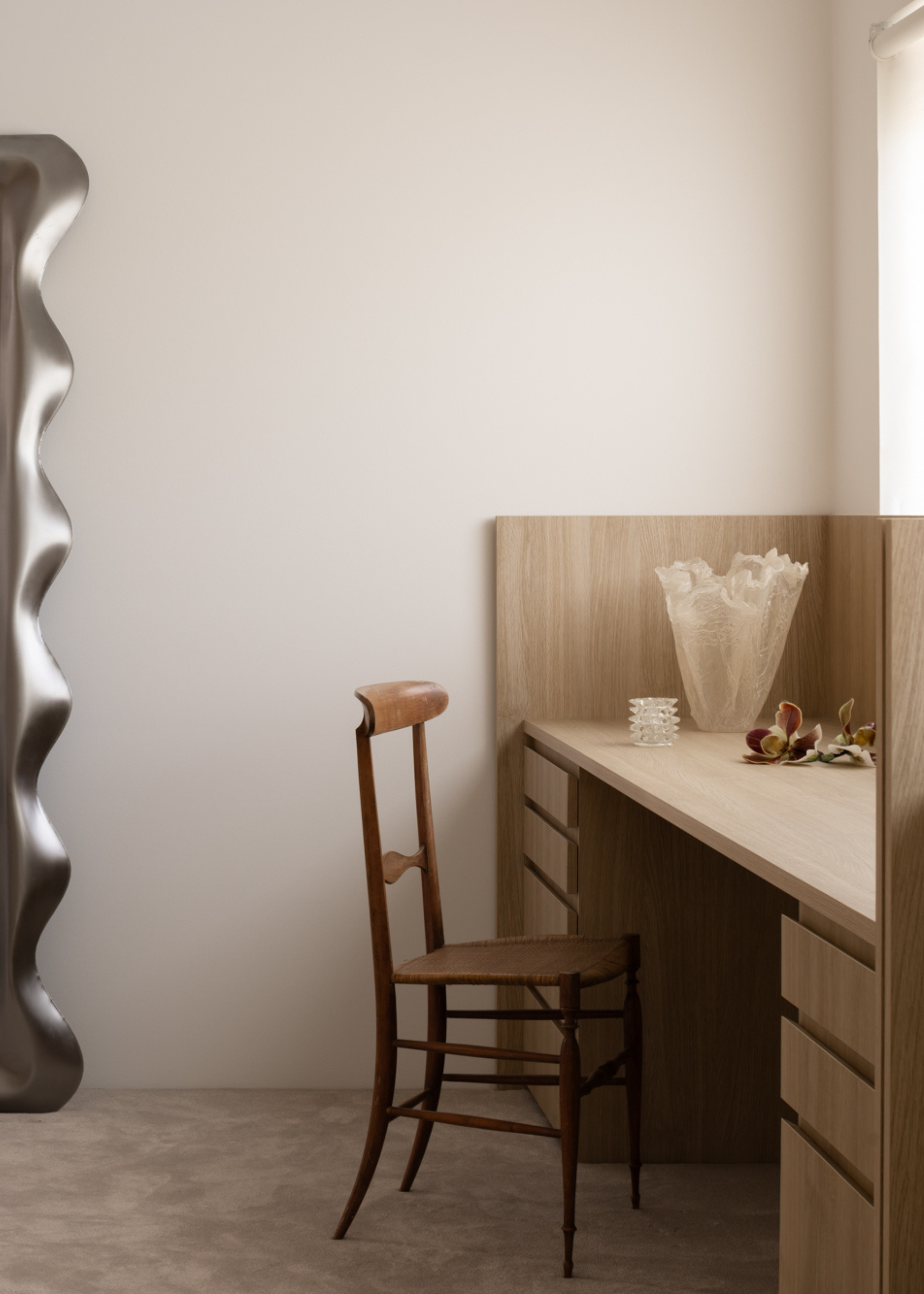 A bedroom with a wood vanity, a glass vase, a wood chair, and a curved brushed chrome mirror