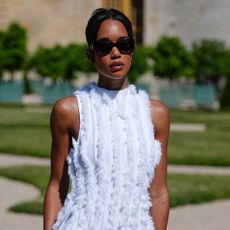 VERSAILLES, FRANCE - JUNE 29: Laura Harrier wears a sleeveless white floor-length dress with textured vertical ruffled stripes and a high neckline, styled with dark burgundy pointed-toe heels and a glossy burgundy croc-embossed leather handbag. Hair is pulled back in a sleek low bun and makeup includes bold brows and a matte lip. Black rectangular sunglasses complete the look, outside Jacquemus, during the Paris Fashion Week - Menswear Spring/Summer 2026, on June 29, 2025 in Versailles, France. 