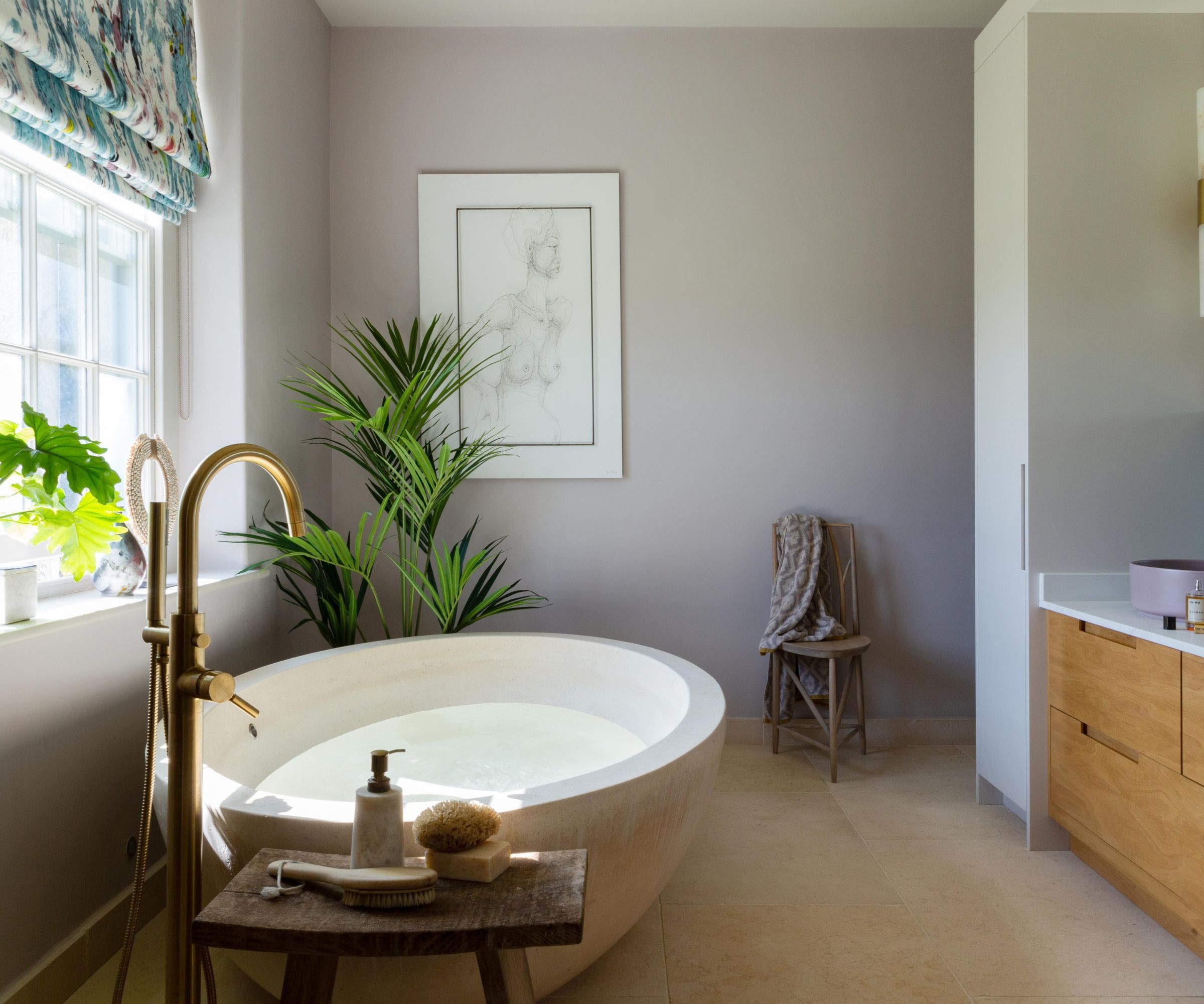 A soft, serene bathroom with lilac-grey walls and a large round stone freestanding bath with a brass floor-mounted tap. A tall indoor palm sits beneath a framed figure sketch, beside a rustic wooden stool with bath accessories. A floral roman blind and oak vanity unit add warmth.