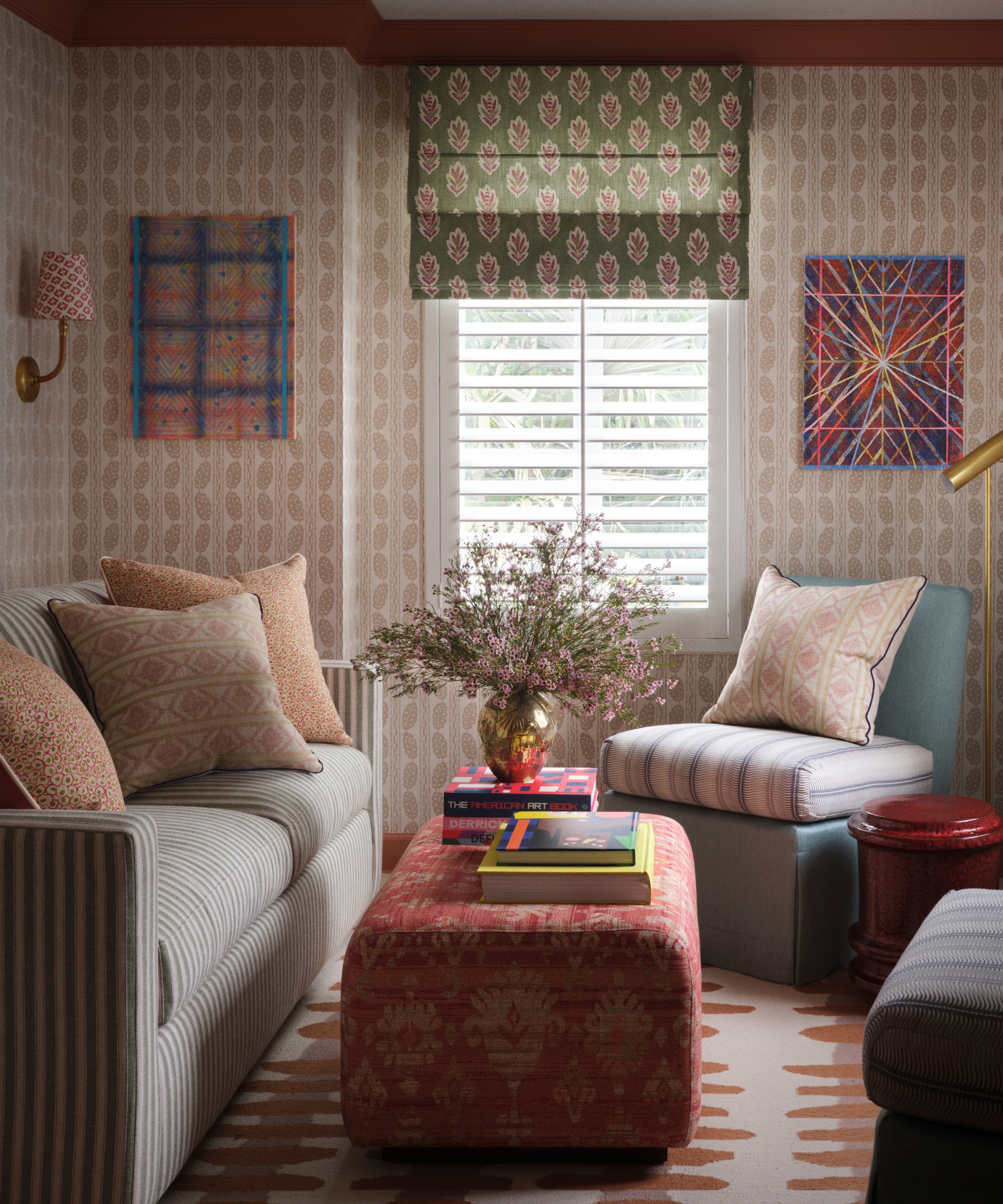 Study with striped sofa, patterned wallpaper, green floral blind, coral ottoman, contemporary art and layered textiles.