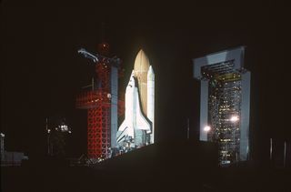 A nighttime view of the space shuttle Enterprise, mated to an external tank and solid rocket boosters, resting on the launch mount next to the access tower at Space Launch Complex Six.