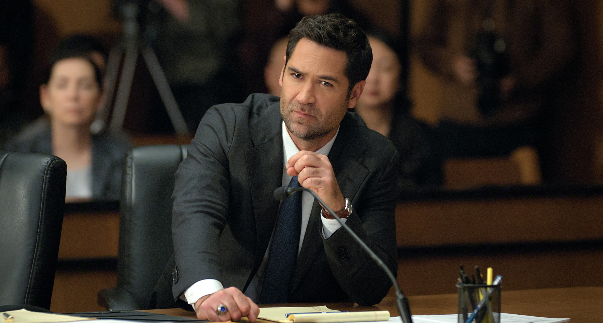 Manuel Garcia-Rulfo as mickey heller wearing a black suit sitting at a table in a court room in the lincoln lawyer season 4