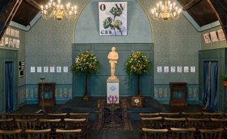 The Oscar Wilde Temple, rows of wooden chairs on either side of an isle leading to an Oscar Wilde cream statue, tall potted flowering plants, dark wooden floor with colourful scatter rugs, arched wooden ceiling, two lit chandeliers, decorated walls with colourful artwork, two doorways with blue and gold curtains