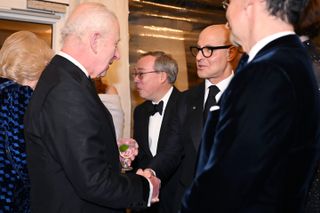 King Charles wearing a tuxedo shaking hands with Stanley Tucci, with Queen Camilla wearing a blue and black dress in the background talking to another man in a tux