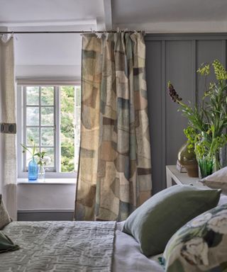 gray and white bedroom with gray panelling, cubist style drapes with neutral pattern, sage green bed pillow, bedspread, vases of flowers