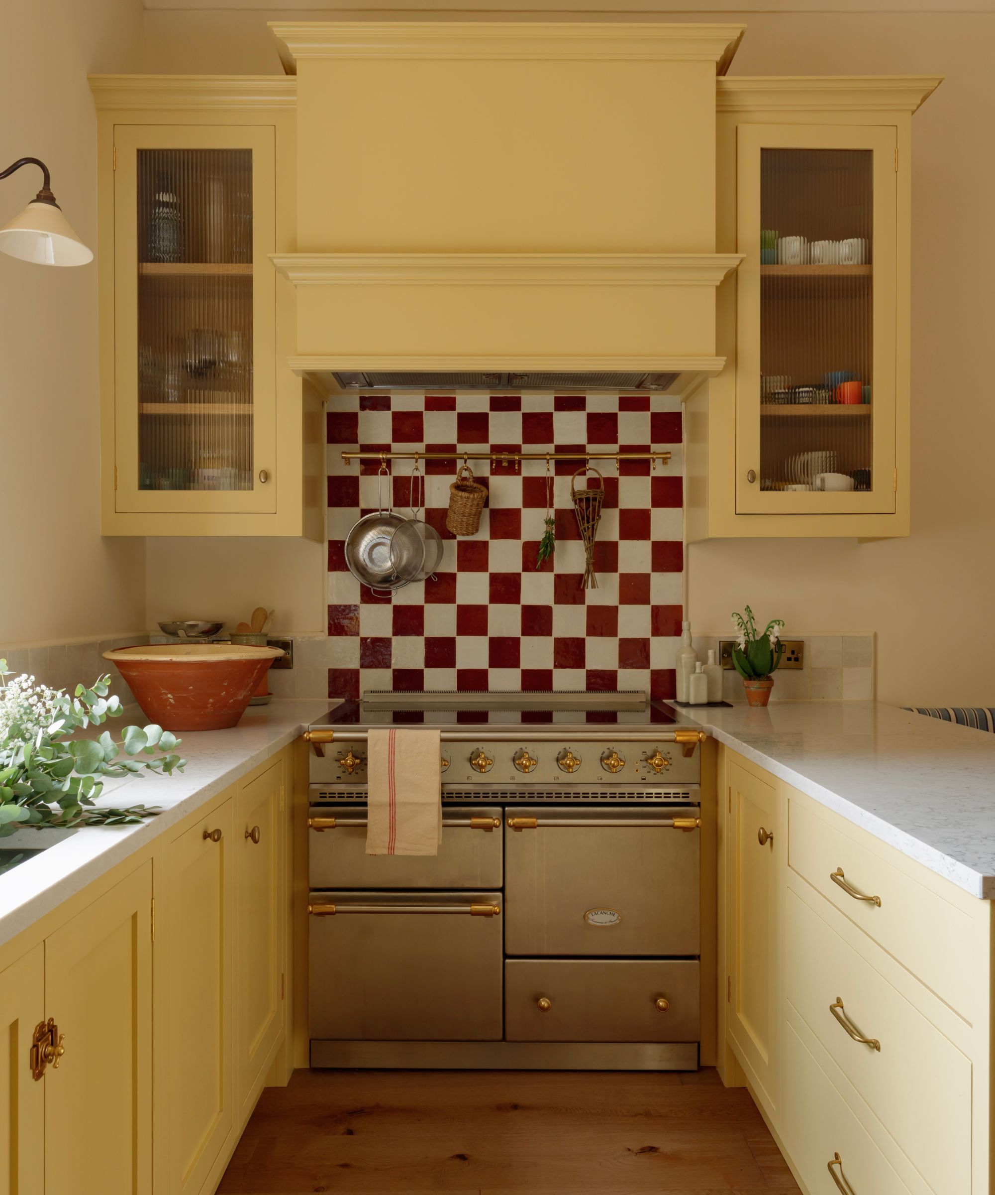Small Georgian kitchen with Shaker cabinets in yellow, red and white checkerboard backsplash, brass range cooker, and tall glazed storage units.
