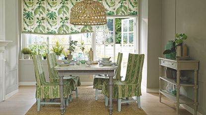 A green green dining room with a jute rug, light wood flooring, and a pale grey table in the centre of the room, surrounded by four green and white patterned chairs. To the right is a white side table, and in the background are large windows with green and white botanical print blinds, with various mismatched plants on the windowsill.
