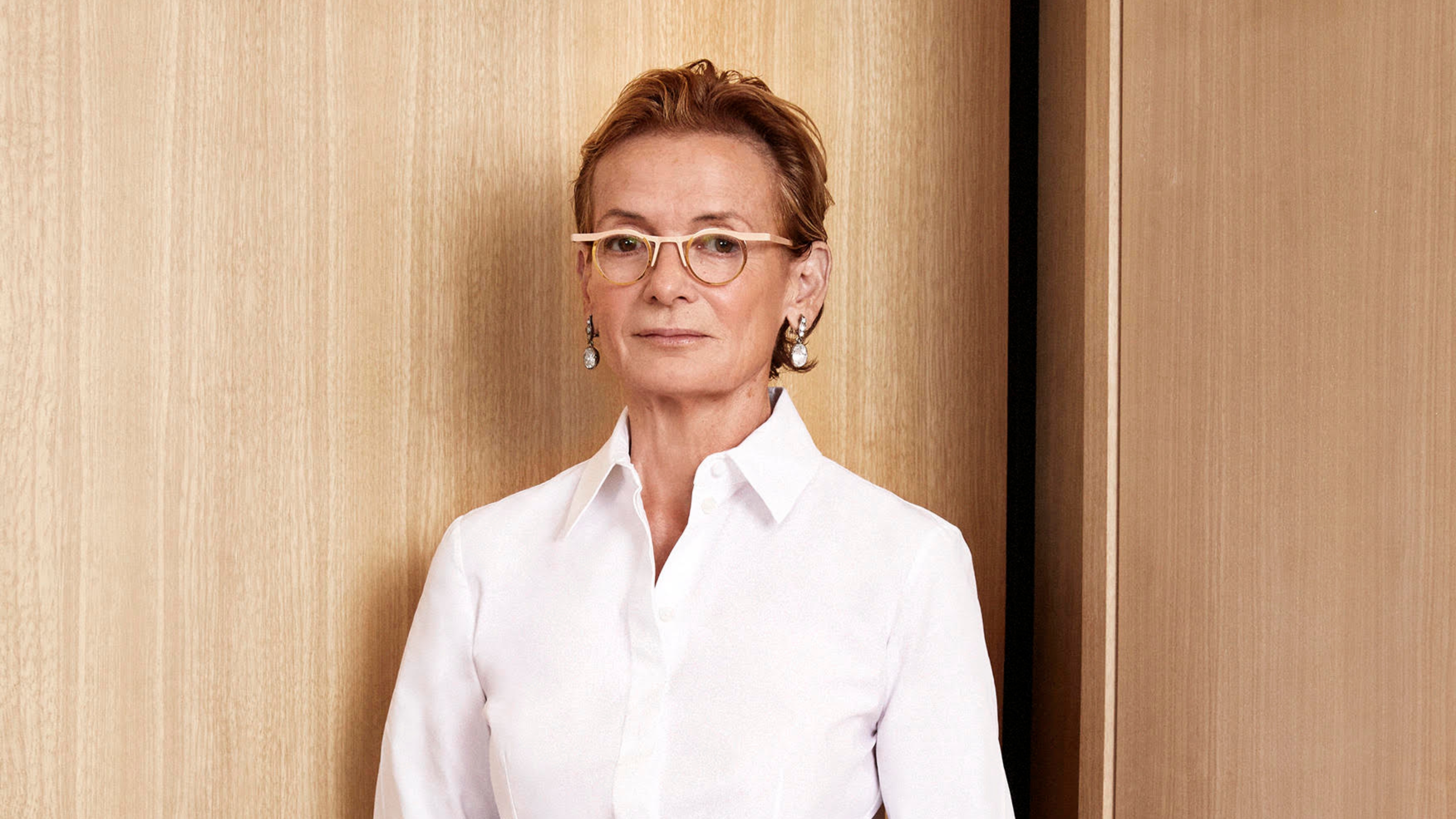 A woman in a smart white shirt, with beige and brass-decorated prescription glasses and wide silver earrings looks at the camera while standing in front of a wooden wall.