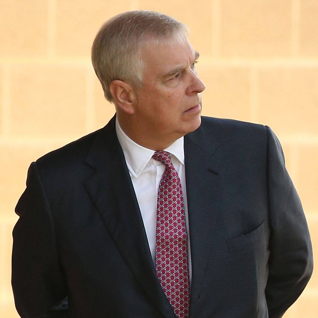 Ex-Prince Andrew wearing a red tie and suit standing in front of a yellow brick wall