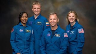 four smiling⁤ people in blue flight suits stand in front of a dark ‍gray-brown background.