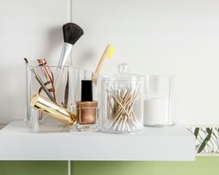 Acrylic makeup organizers on shelf with green tiling behind with cotton buds, makeup, cotton pads, brushes and eyelash curlers