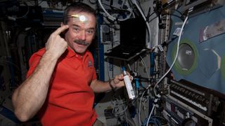 an astronaut pointing at a skin sensor on his forehead while floating in a space station module