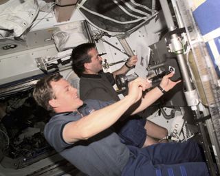 Astronaut Chris Hadfield, far right, pictured working in close quarters with a colleague on board a space shuttle during a mission. They are floating in zero gravity, and surrounded by equipment including wired, tubing, metalwork