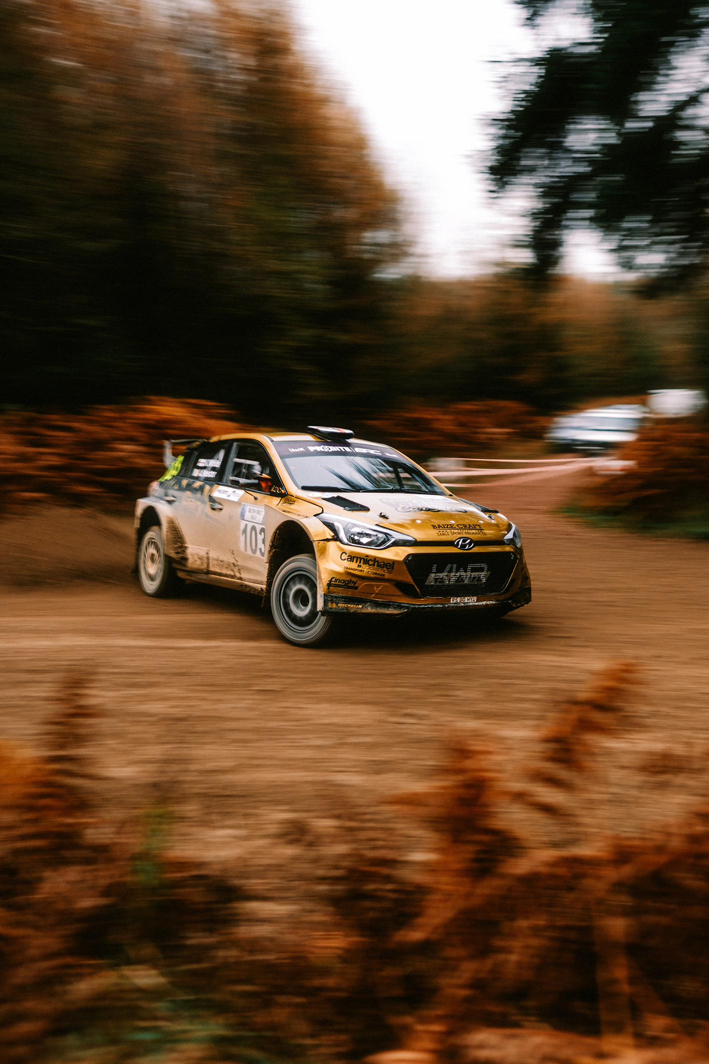 A golden rally car speeds around a dirt track, surrounded by autumn foliage, with motion blur emphasizing its dynamic movement